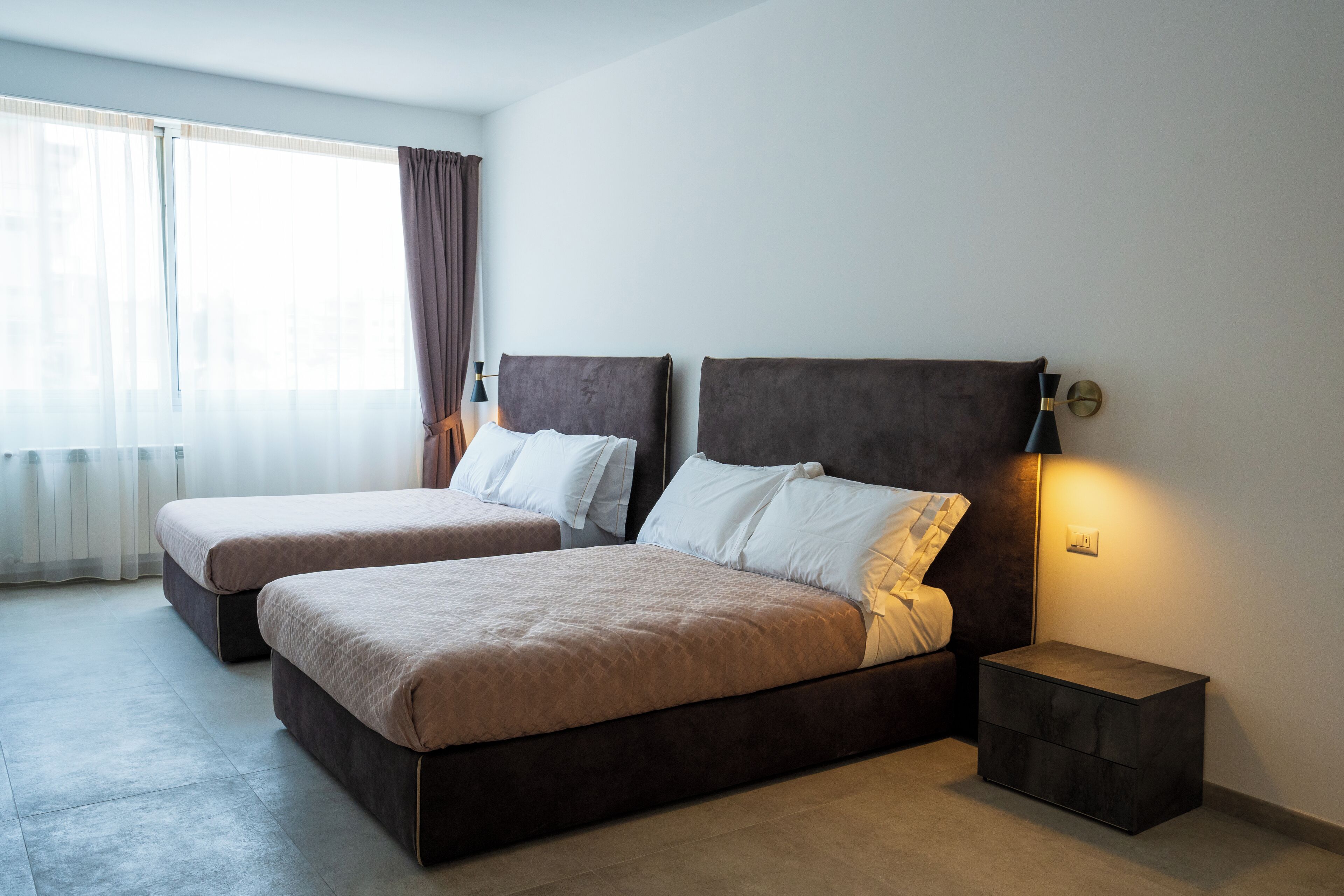 Hotellihuone, jossa on kaksi sänkyä, yöpöytä, lamppu ja ikkuna, jossa on verhot.