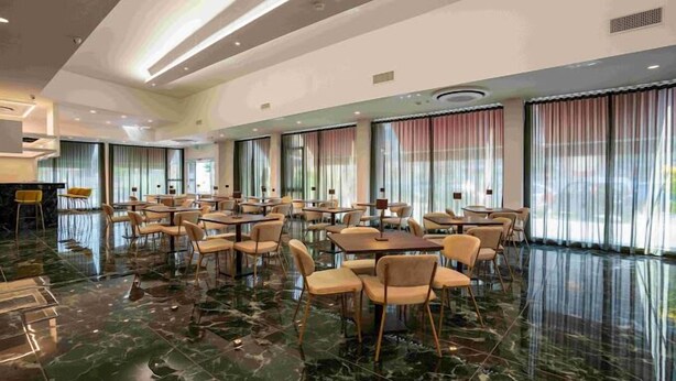 A spacious dining area with marble flooring, large windows with curtains, and multiple tables and chairs.