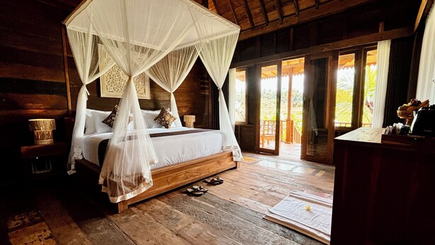 A wooden four-poster bed with a canopy, a nightstand with a lamp, and a view of a garden through a glass door. - Jagadhita Villas Ubud Bali (Ubud)