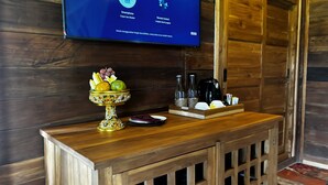 A wooden console table with a fruit bowl, a coffee maker, and a remote control on a wall-mounted TV displaying text in a foreign language. - Jagadhita Villas Ubud Bali (Ubud)