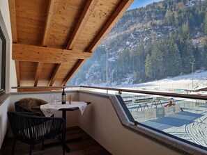A balcony with a wooden ceiling, a table with a wine bottle, and a chair. A snowy mountain landscape is visible through the window. - Nice apartment for 8 guests with WIFI and terrace (Mitterdorf)