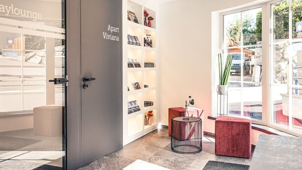 En modern inredning med en grå dörr med texten ”Apart Viviana”, en glasdörr som leder ut, och en röd, vadderad bänk.