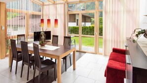 Un espace repas moderne avec une grande table, des chaises et un patio vitré offrant une vue sur une maison et un jardin.