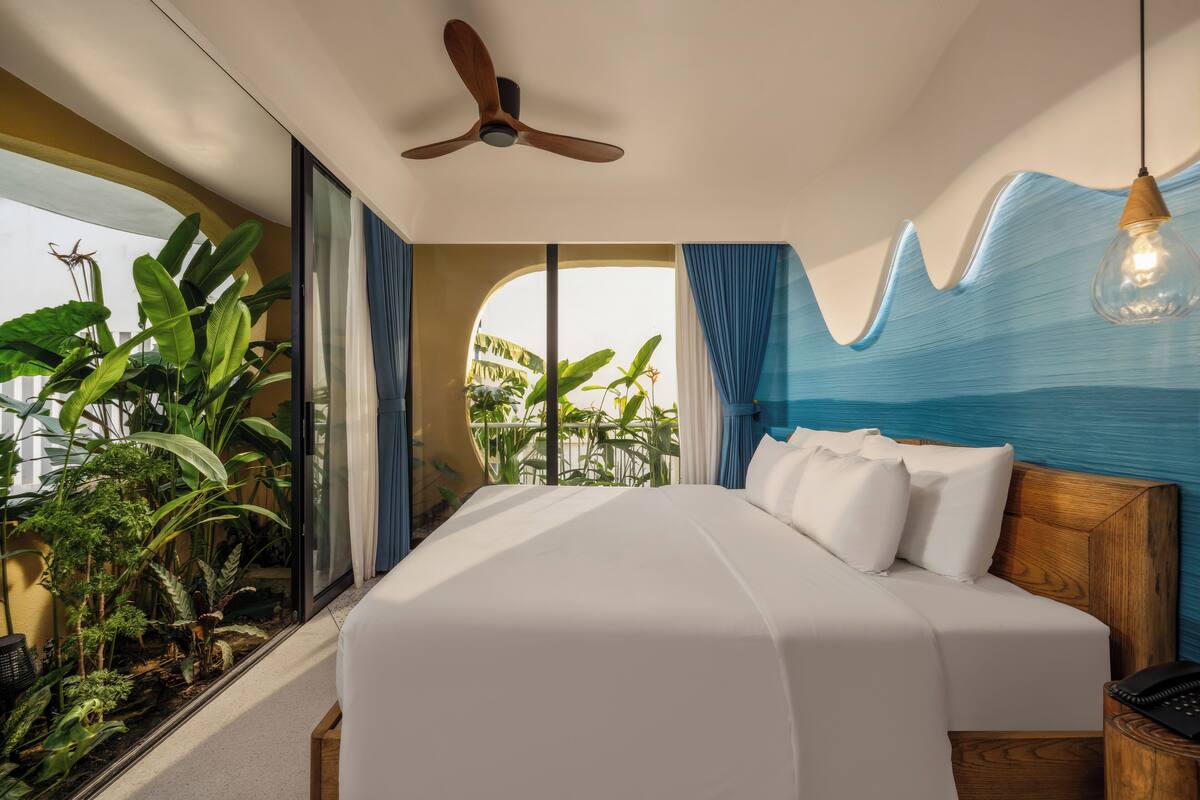 A bedroom with a bed, a ceiling fan, a view of greenery, and a blue wall feature.
