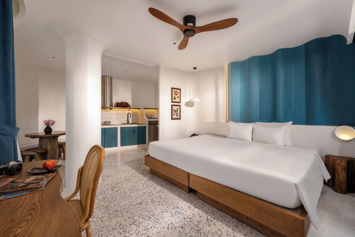 A hotel room with a large bed, wooden headboard, a ceiling fan, a kitchenette, and a dining area with a table and chair.