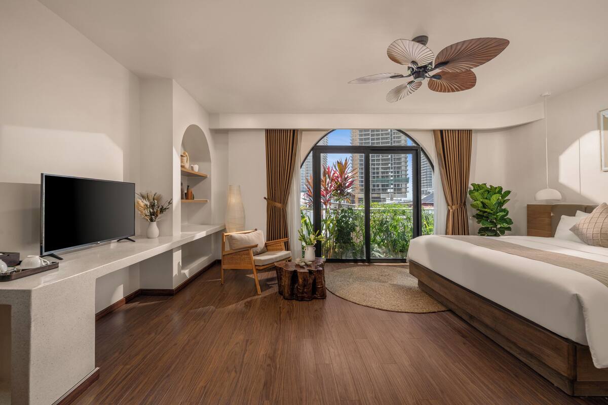 A modern hotel room with a flat-screen TV, a wooden coffee table, a comfortable chair, a large bed, and a view of the city through floor-to-ceiling windows.