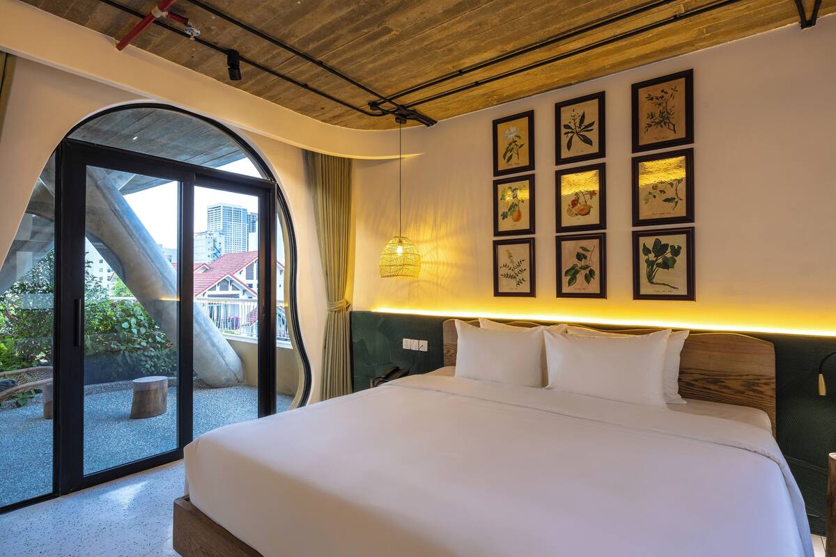 A modern hotel room with a large bed, a city view through a glass door, and framed botanical artwork on the wall.