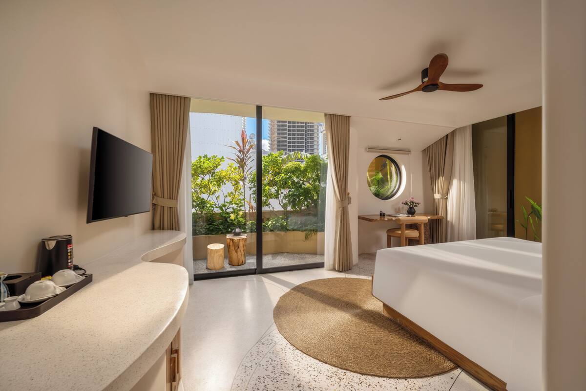 A hotel room with a large sliding glass door to a balcony, a flat-screen TV, a round mirror, a bed, a desk with a chair, and a ceiling fan.