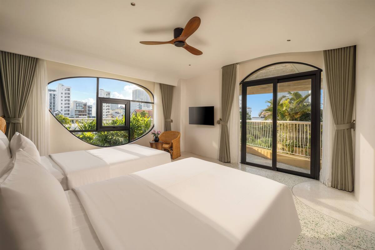 A hotel room with a large bed, a ceiling fan, a TV, a window with a view of buildings and palm trees, and a balcony with curtains.