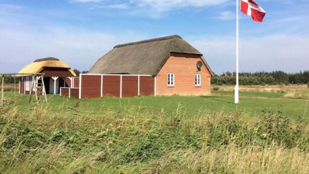 Una casa tradicional de rajola vermella amb un teulat de palla, una bandera blanca i una petita construcció groga al fons.