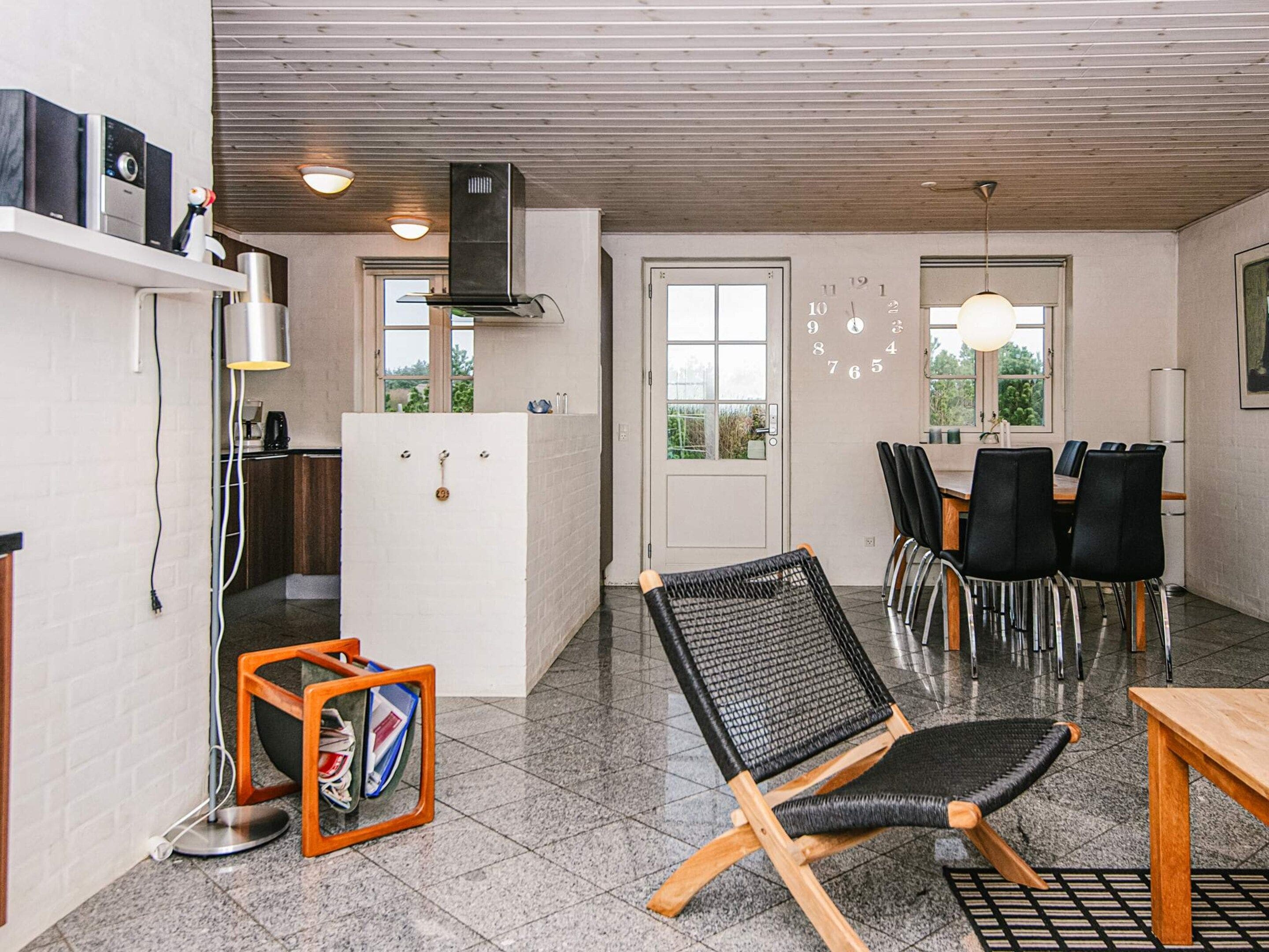 A modern kitchen with a built-in oven, a refrigerator, and a coffee machine. There is a dining area with a table and chairs, and a large window providing natural light.