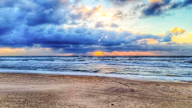 Een strand met golven, een bewolkte lucht en een ondergaande zon.
