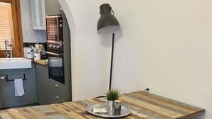 Un coin repas avec une table en bois rustique, des chaises noires, un lampadaire et une cuisine en arrière-plan.