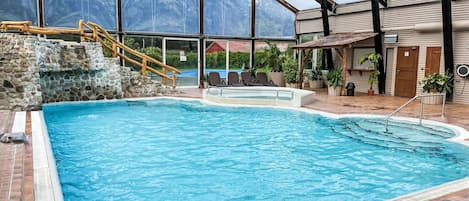 An indoor swimming pool with a stone wall, wooden deck, and a covered area with chairs and a fan.