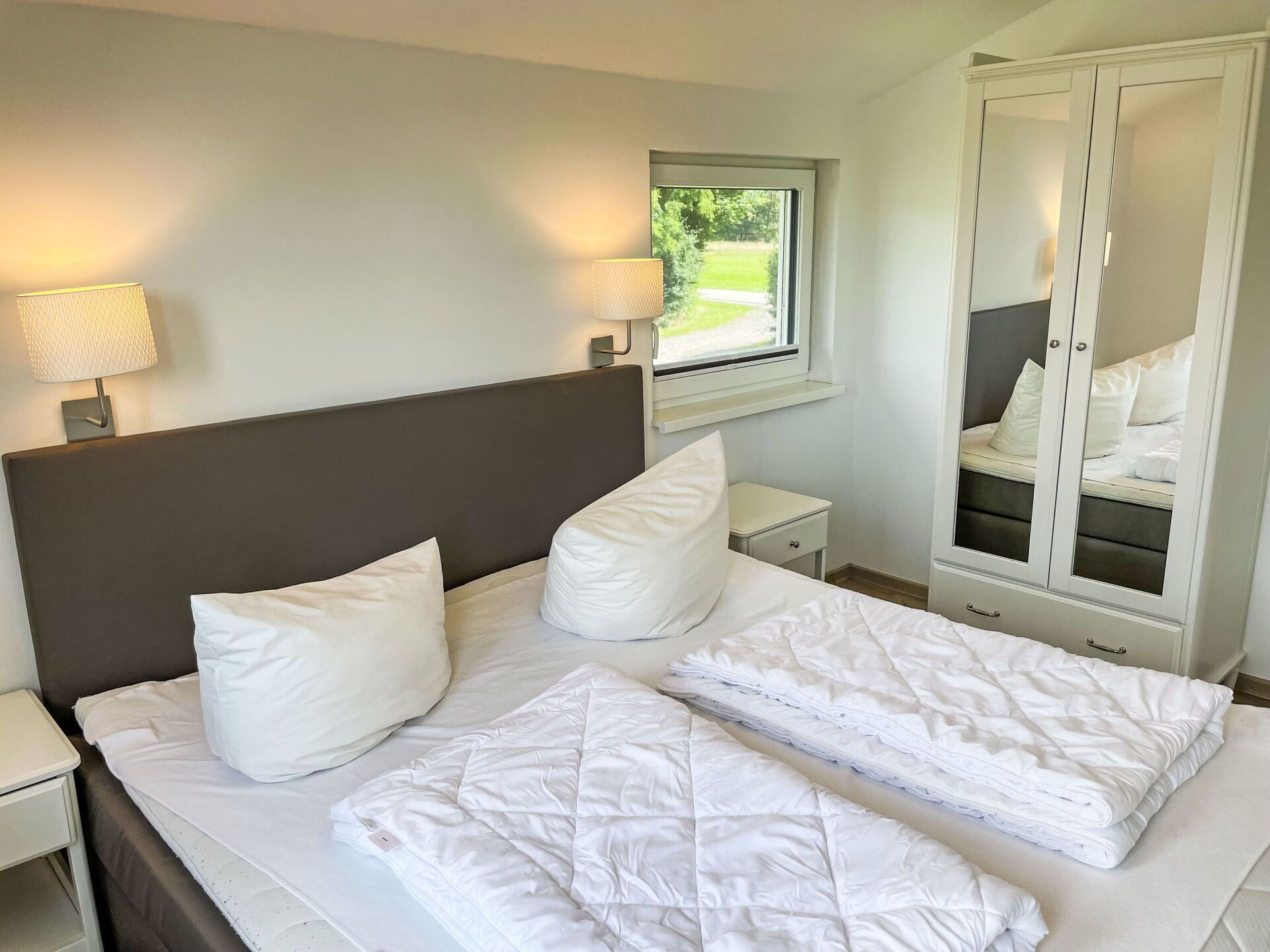 Een keurig opgemaakt bed met witte lakens, een bedlampje, een nachtkastje, een raam met uitzicht op groen, en een garderobe met spiegeldeuren.