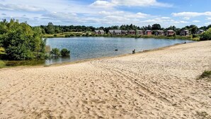 Una playa arenosa con huellas, un lago tranquilo y casas al fondo.