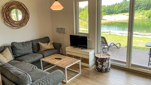 Un salón moderno con un sofá gris, una mesa de centro de madera, un televisor sobre un mueble blanco y un ventanal grande con vistas a un lago.