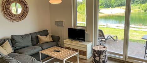 Een moderne woonkamer met een grijze bank, een houten salontafel, een televisie op een witte dressoir en een groot raam met uitzicht op een meer.