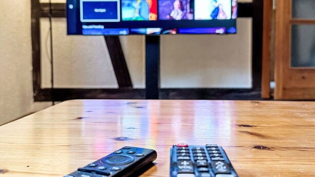 Een televisie toont een startscherm met verschillende app-icoontjes, twee afstandsbedieningen op een houten tafel, en een lamp op de achtergrond.