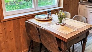 Een houten eettafel met stoelen, een raam met uitzicht op groen en een klein kastje met een klok.