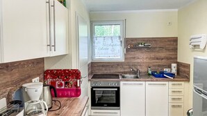 Une cuisine compacte avec des meubles blancs, un four encastré, un évier et une fenêtre donnant sur un espace verdoyant.