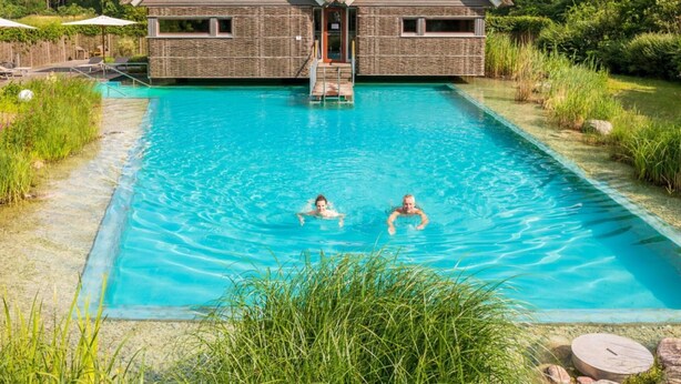 Une piscine où se trouvent deux personnes, entourée d’une maison à toit de chaume et aux murs en pierre.