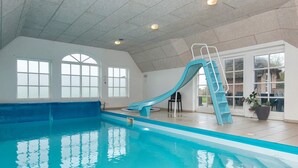 Una piscina interna con uno scivolo blu, ampie finestre e un soffitto piastrellato.
