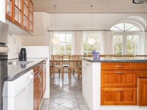 A kitchen with a granite countertop, wooden cabinets, a coffee maker, and a dining area with wooden chairs and a table. - 4 Star Holiday Home in Ulfborg (Ulfborg)