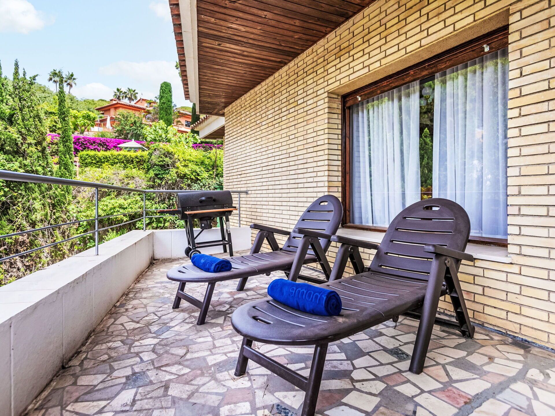 A balcony with two lounge chairs, a grill, and a window with curtains.