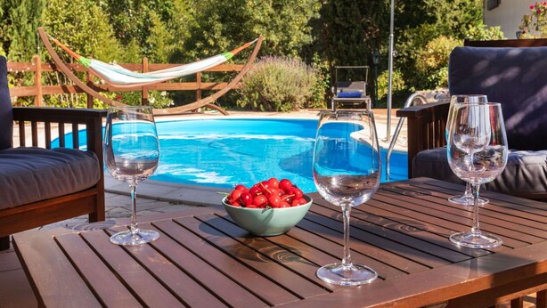 A wooden table with three wine glasses and a bowl of cherries by a pool.