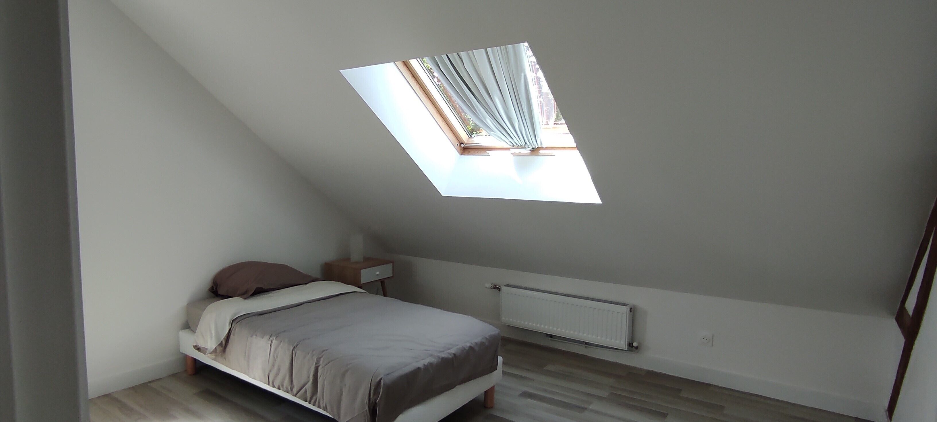 Un dormitorio con techo inclinado, una cama, una mesita de noche, un radiador y una ventana con cortinas.