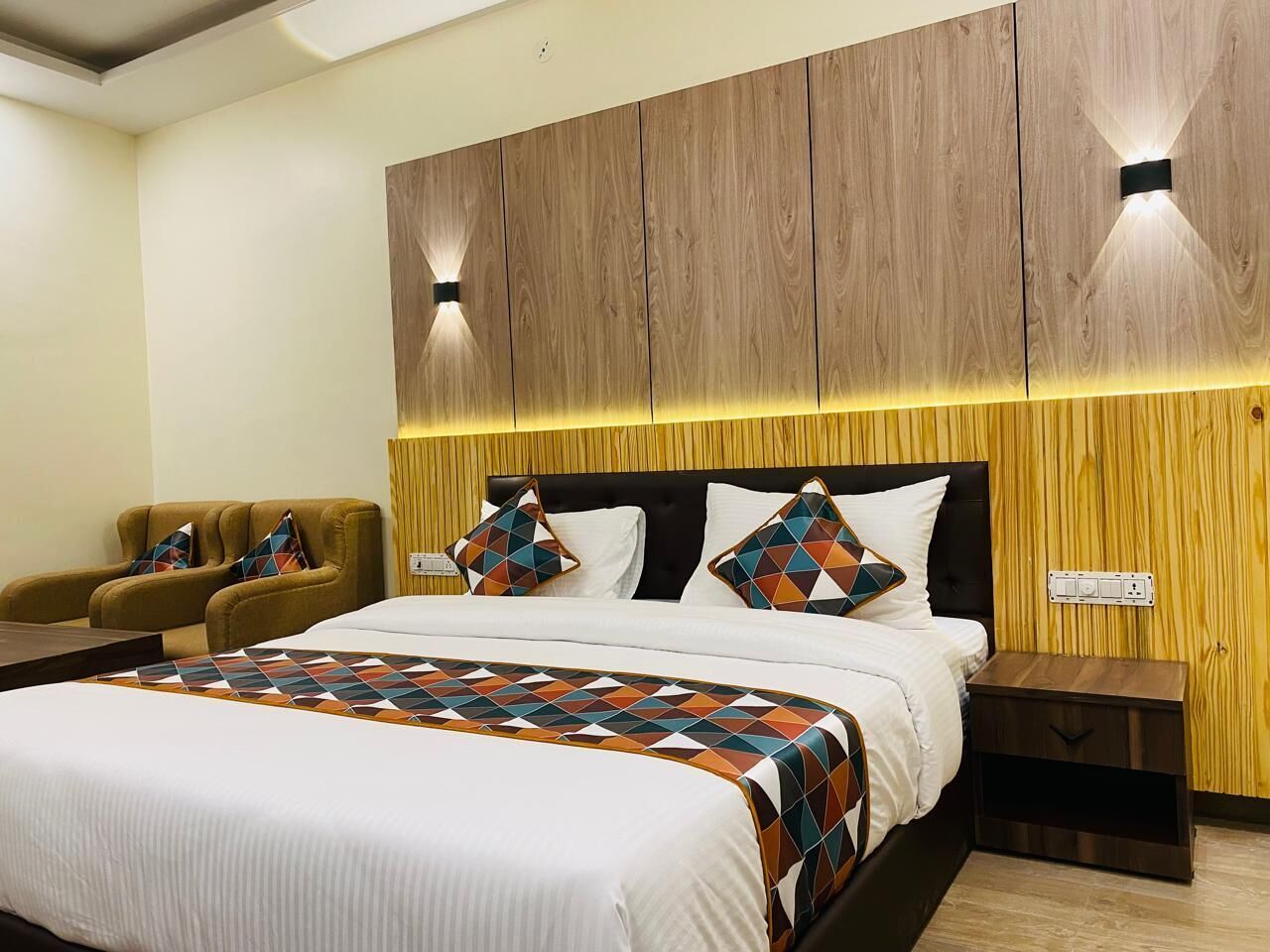 A modern bedroom with a large bed, a wooden headboard, two wall-mounted lights, a nightstand, and a pair of armchairs.