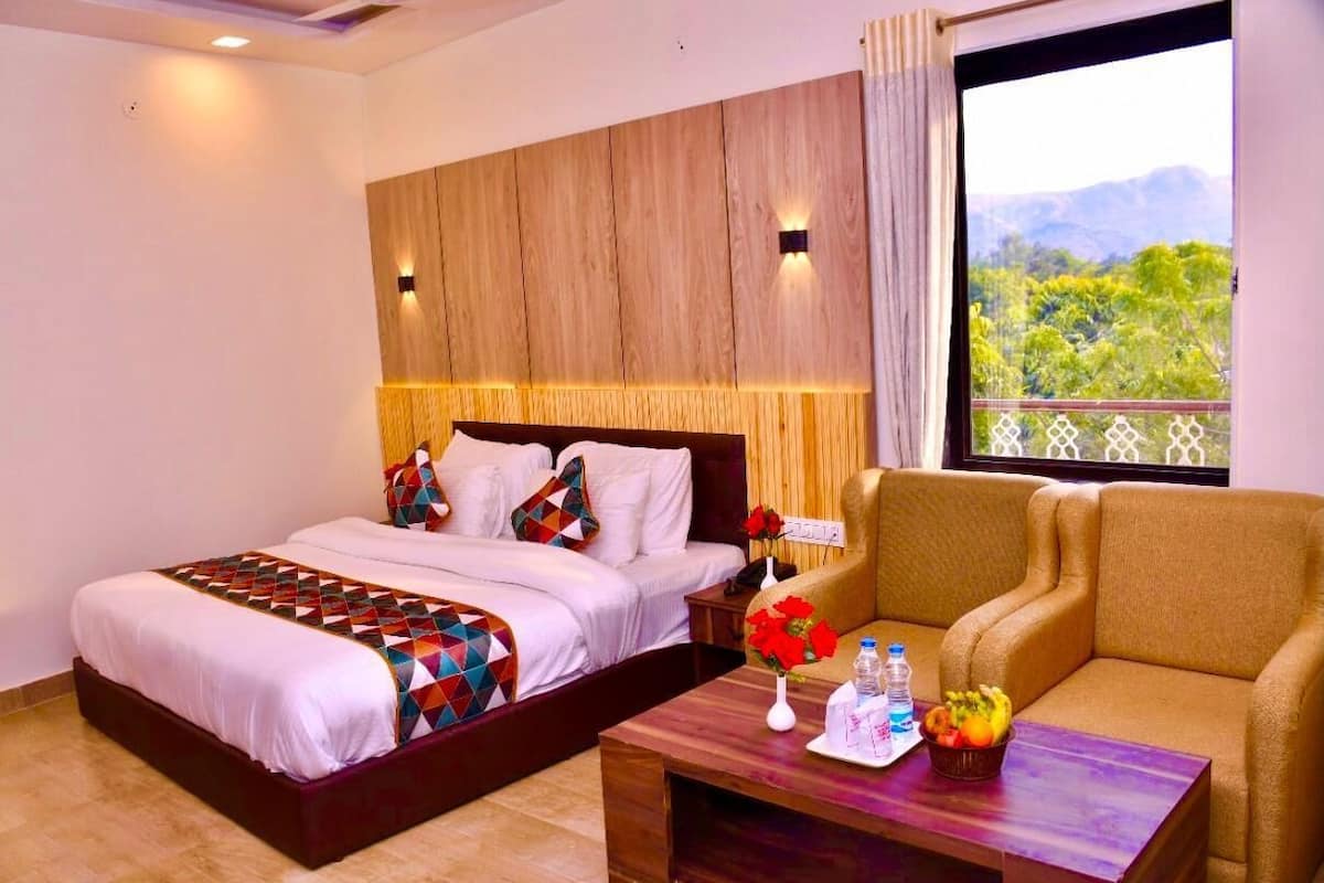 A hotel room with a bed, a sofa, a wooden headboard, a nightstand, a vase with flowers, and a window with a view of greenery and mountains.