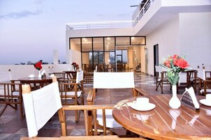 Un área de comedor al aire libre con mesas y sillas de madera, un comedor con paredes de vidrio al fondo y vistas del horizonte de la ciudad.