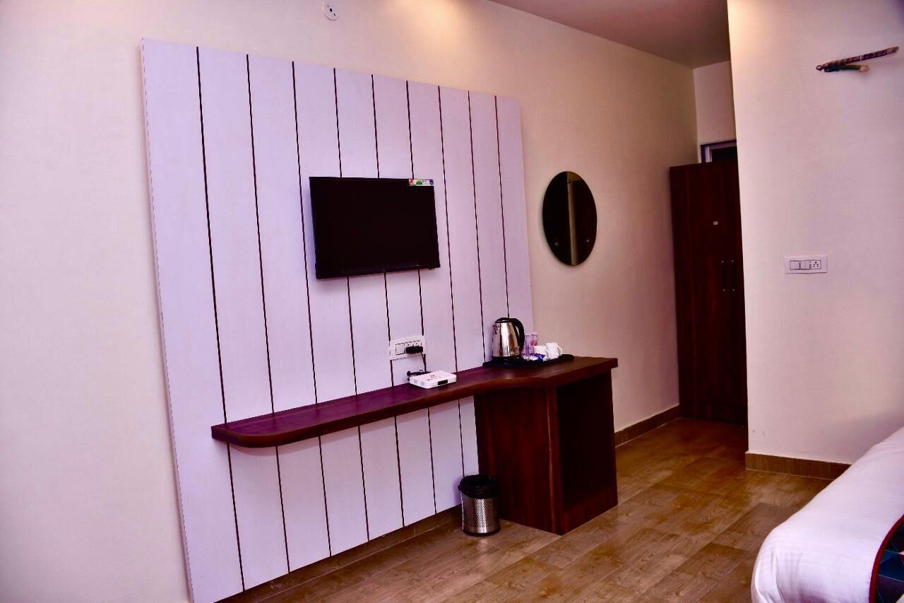 A hotel room with a wooden floor, a TV mounted on a white panel wall, a small wooden desk with a kettle, and a trash bin.