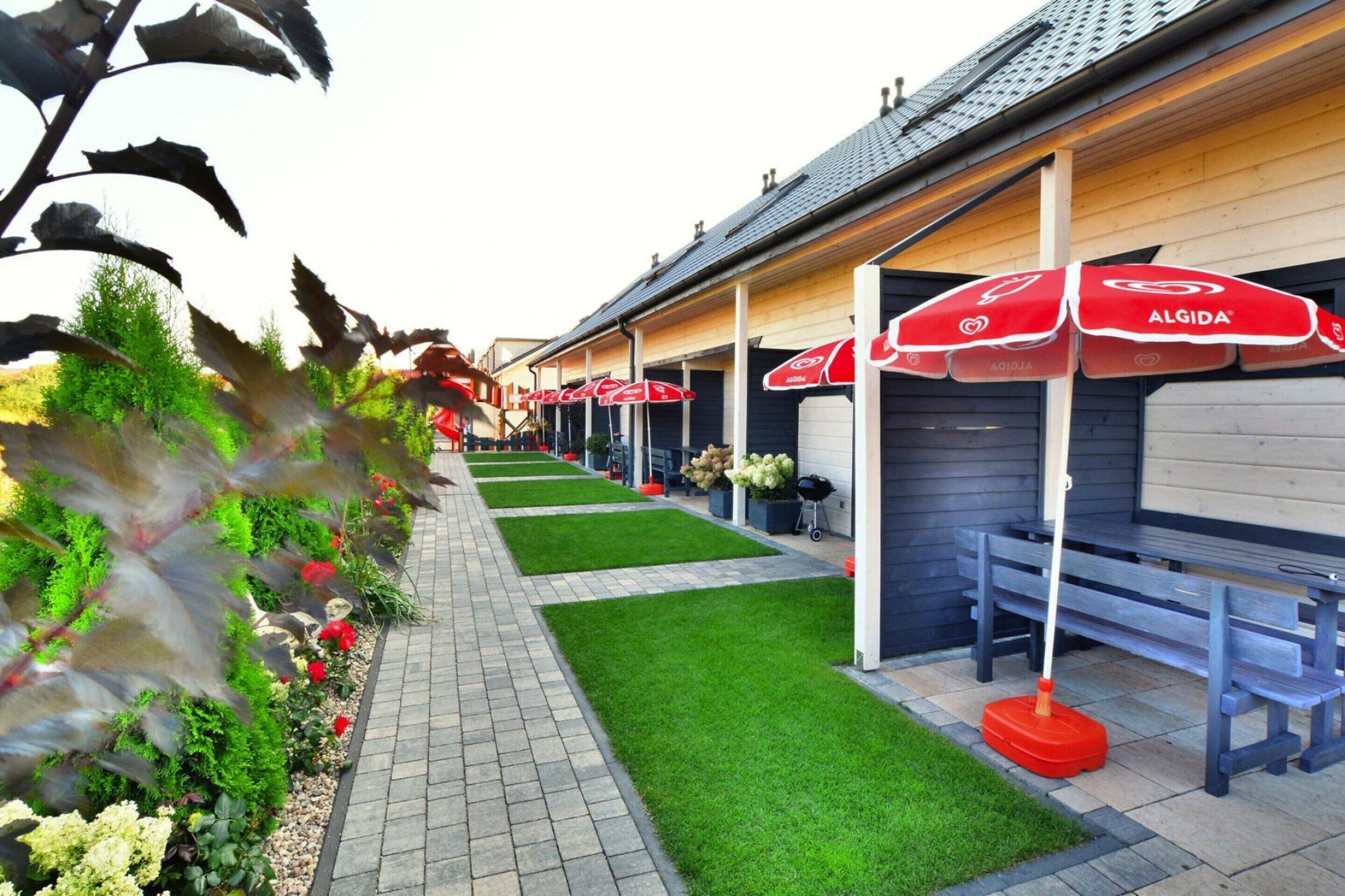 A row of outdoor seating areas with red umbrellas, a paved walkway, and a well-maintained garden with various plants and flowers.