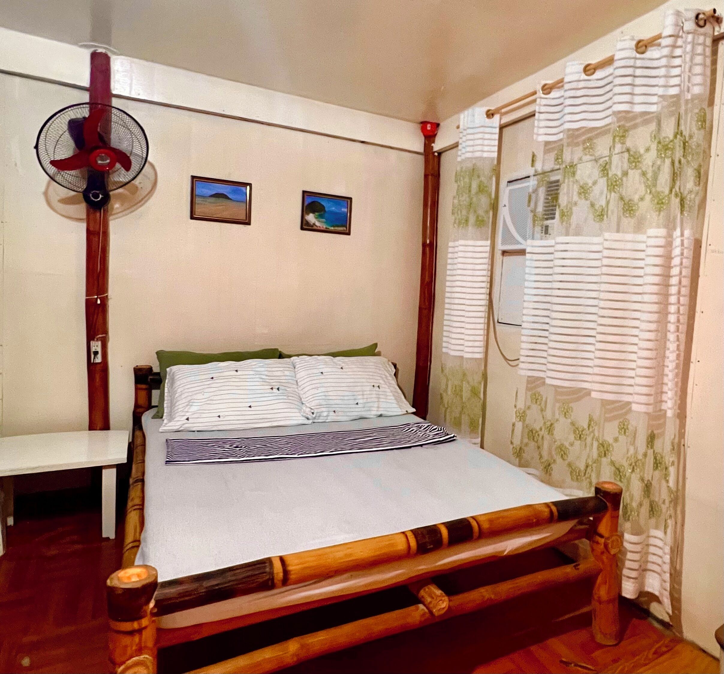 A bedroom with a wooden bed frame, a bedside table, a standing fan, and two framed pictures on the wall.