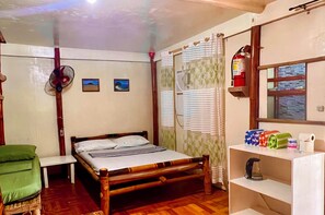 A single bed with a wooden frame, a bedside table, a green chair, a fan, a fire extinguisher, and a window with blinds. - Treasure Beach Co.  (Carles)