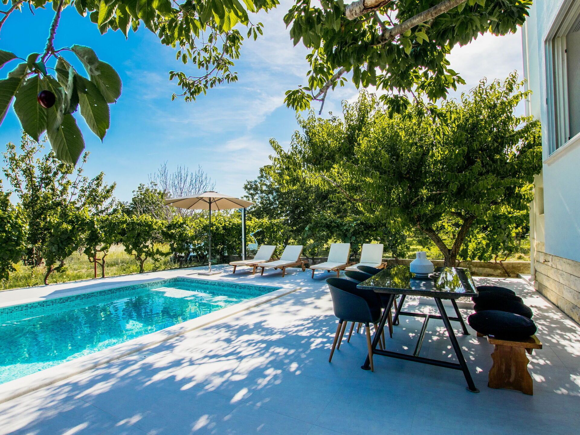Piscina com espreguiçadeiras e uma mesa, cercada por árvores e um edifício.