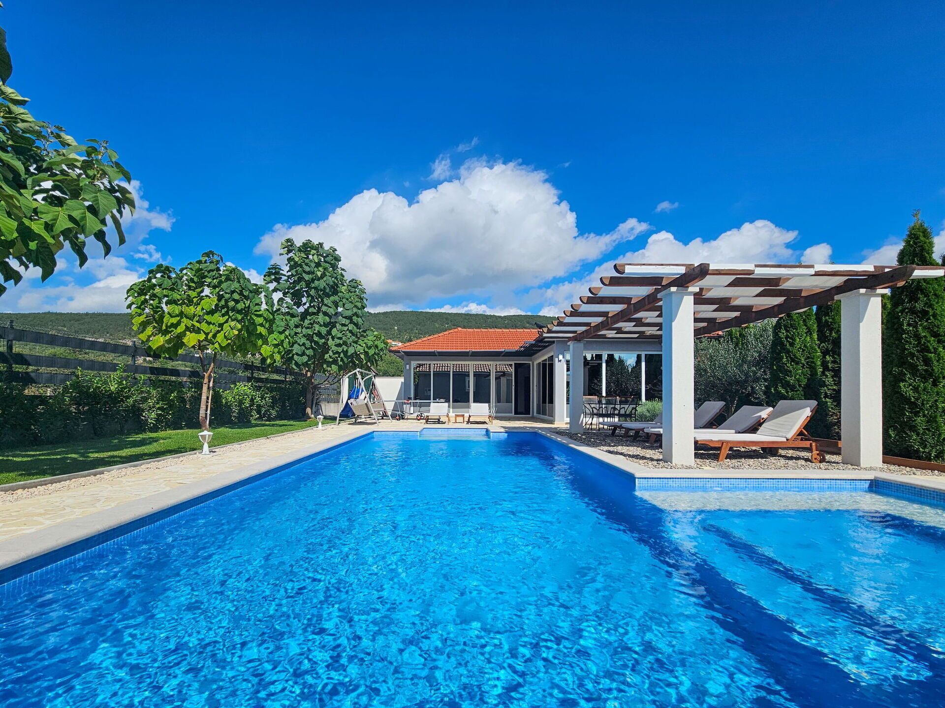 A swimming pool with a pergola and lounge chairs, surrounded by greenery and a clear sky.
