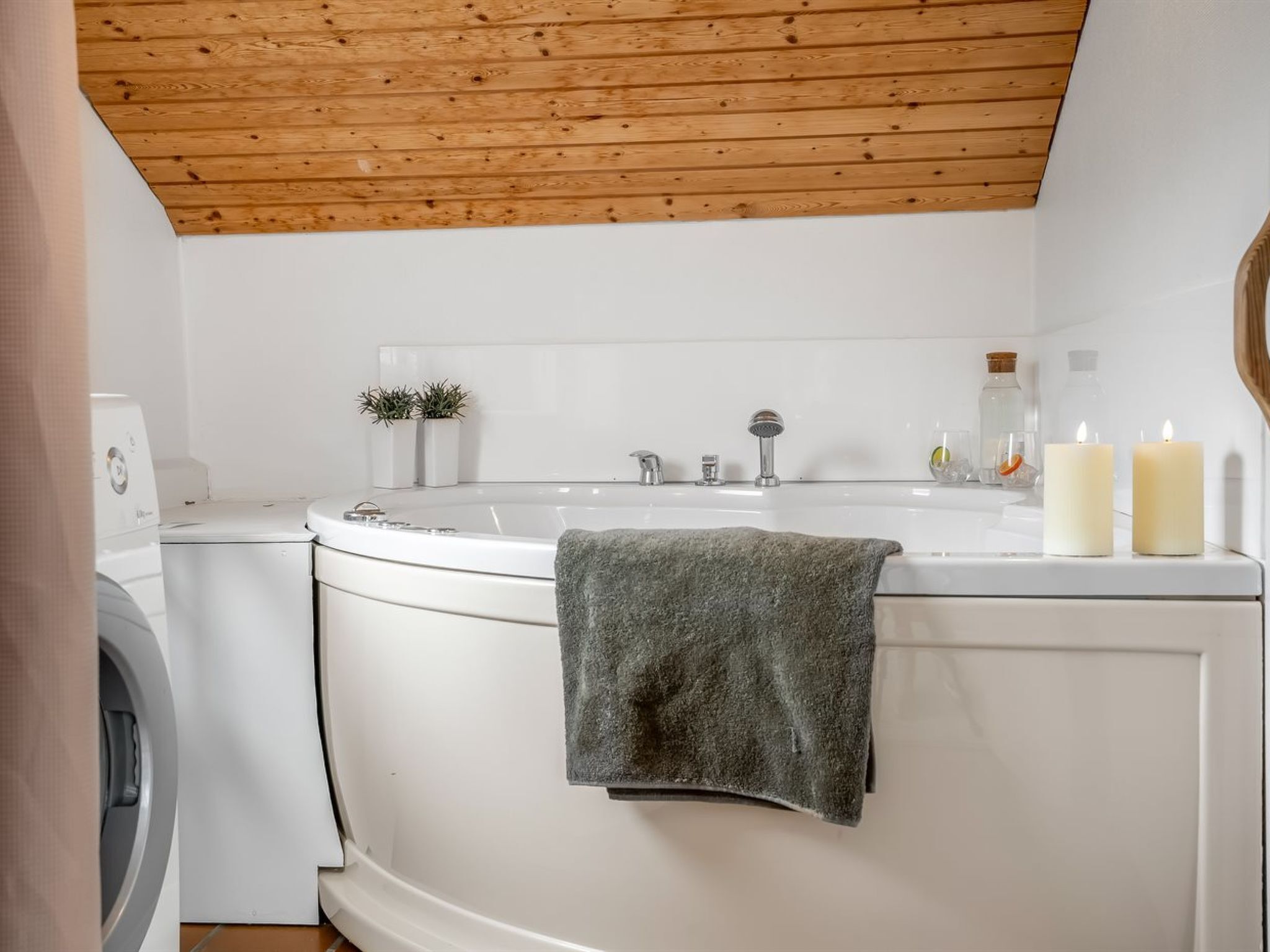 Un baño moderno con techo de madera, lavadora integrada, bañera y dos velas sobre el lavabo.