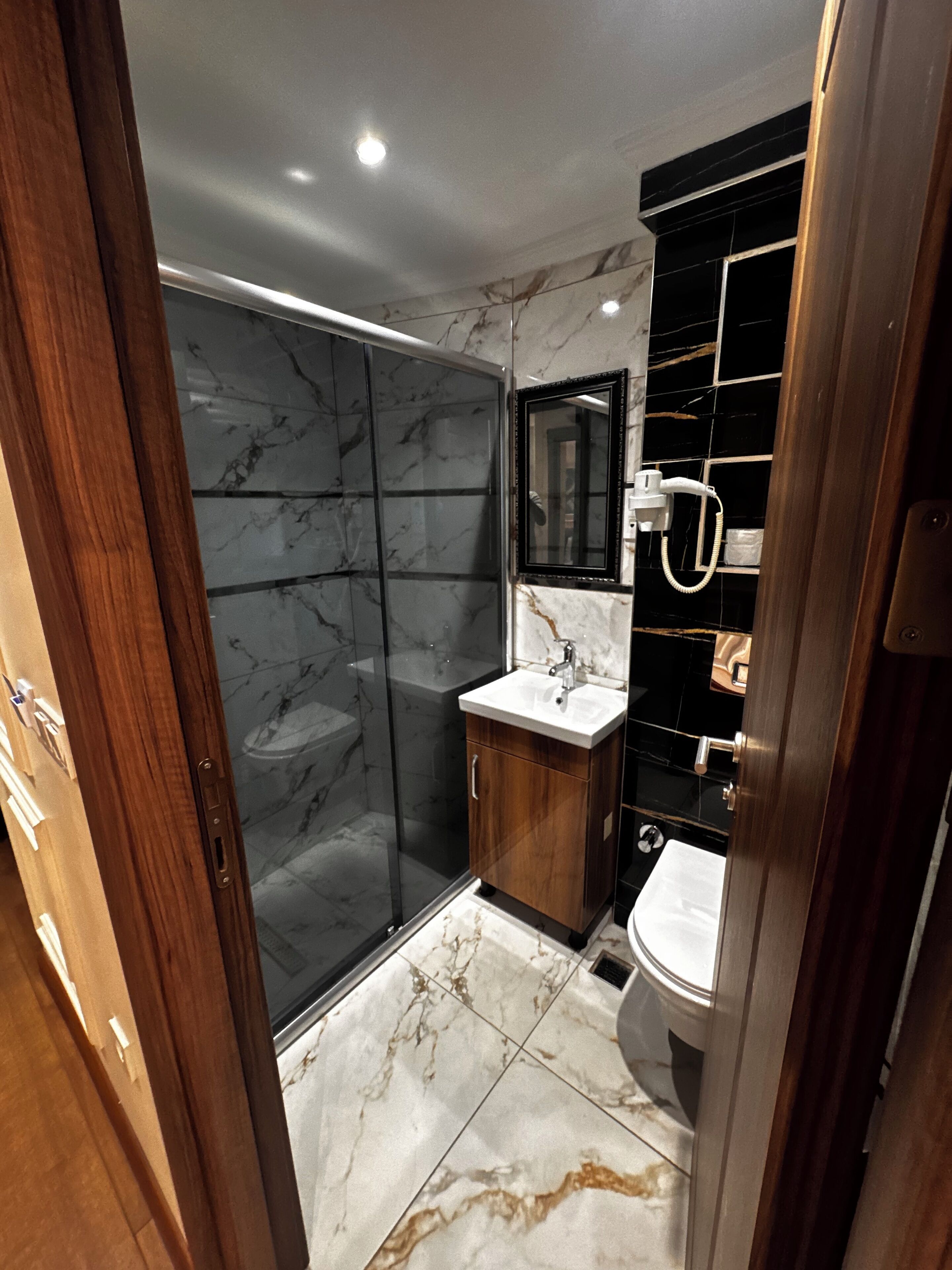 A modern bathroom with a glass shower enclosure, a wooden vanity, and a marble-tiled floor.