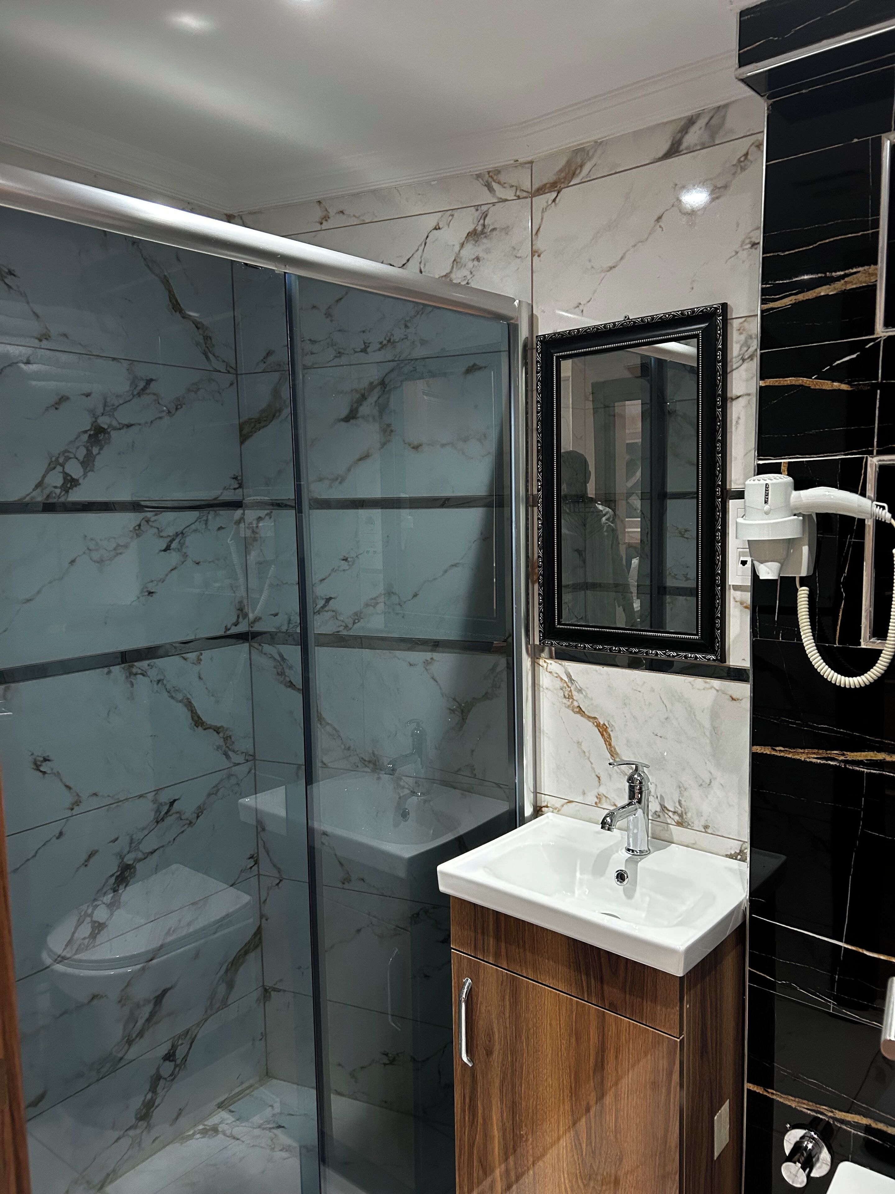 A modern bathroom with a glass shower enclosure, a wooden vanity with a white sink, and a black tiled wall with a mirror.