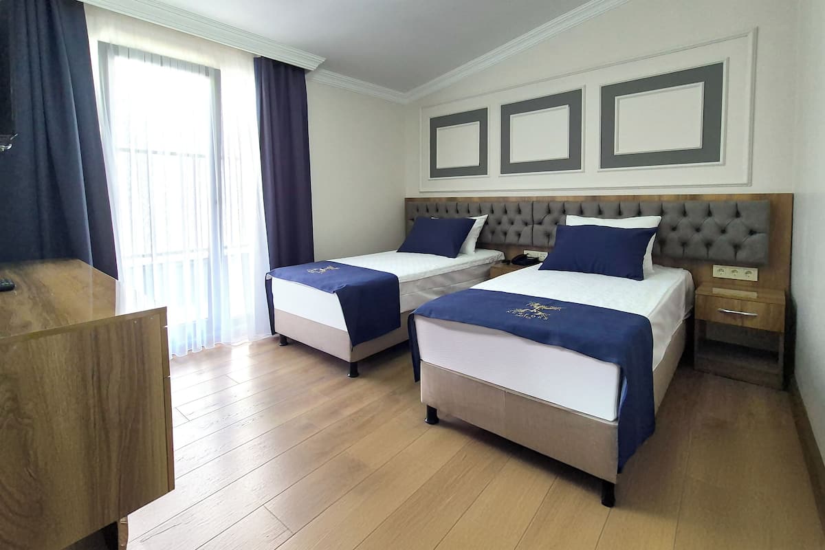 A hotel room with two beds, wooden flooring, a desk, and a window with curtains.
