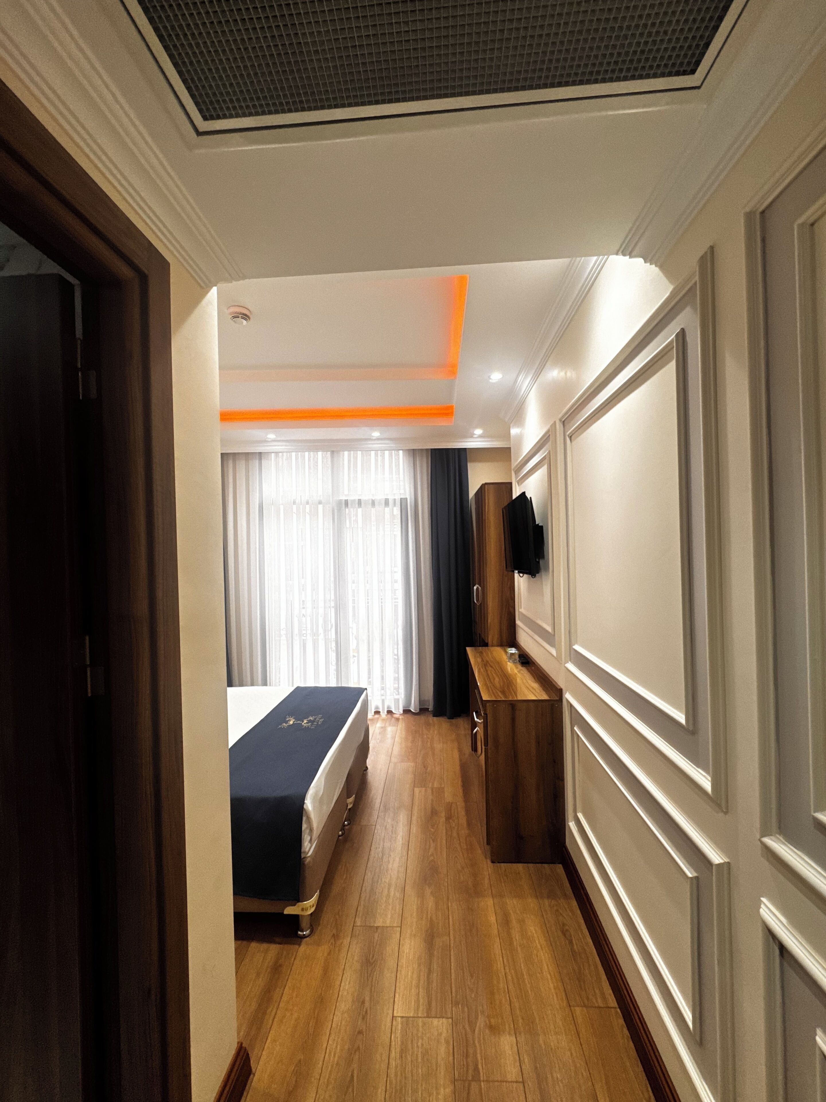 A hotel room with a wooden floor, a bed with a dark cover, a wooden dresser, a flat-screen TV mounted on the wall, and a window with sheer curtains.