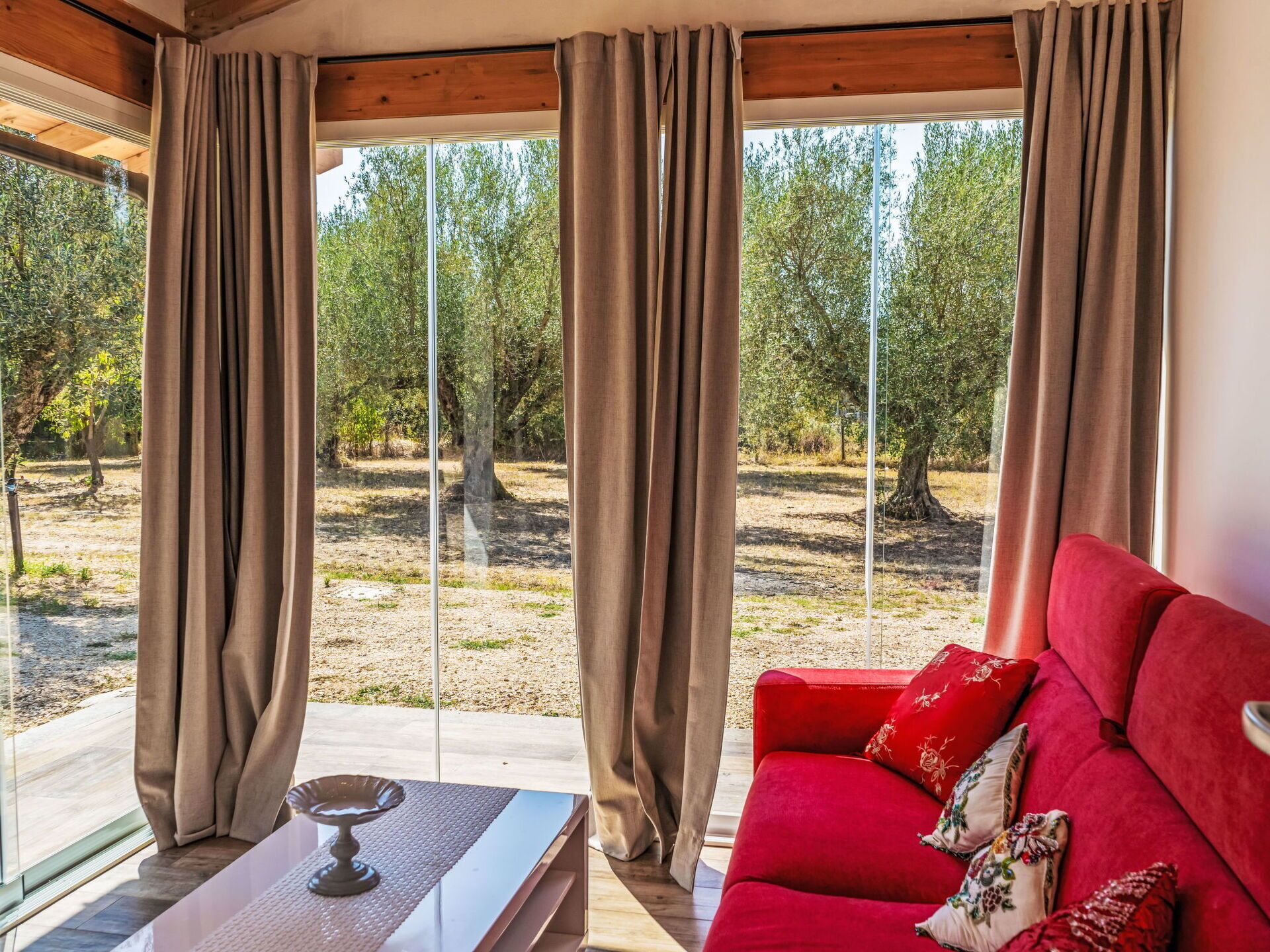 A room with a red sofa, a small table, and a view of trees through large windows with beige curtains.