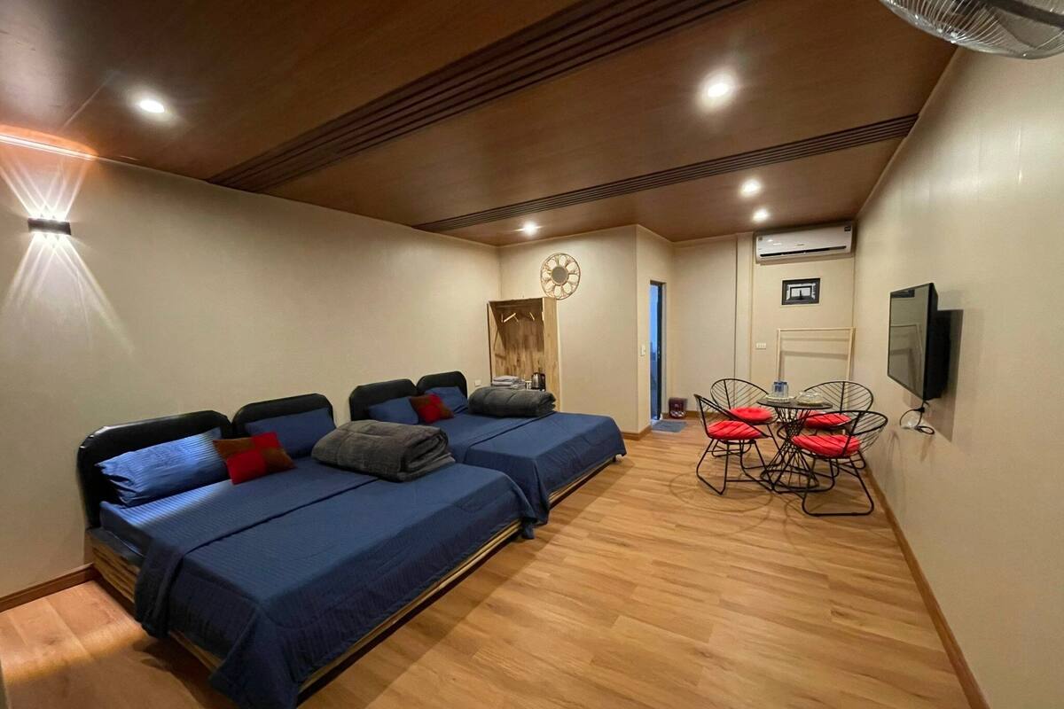 Une chambre avec deux lits, un coin repas avec des chaises, une télévision et une horloge au mur.