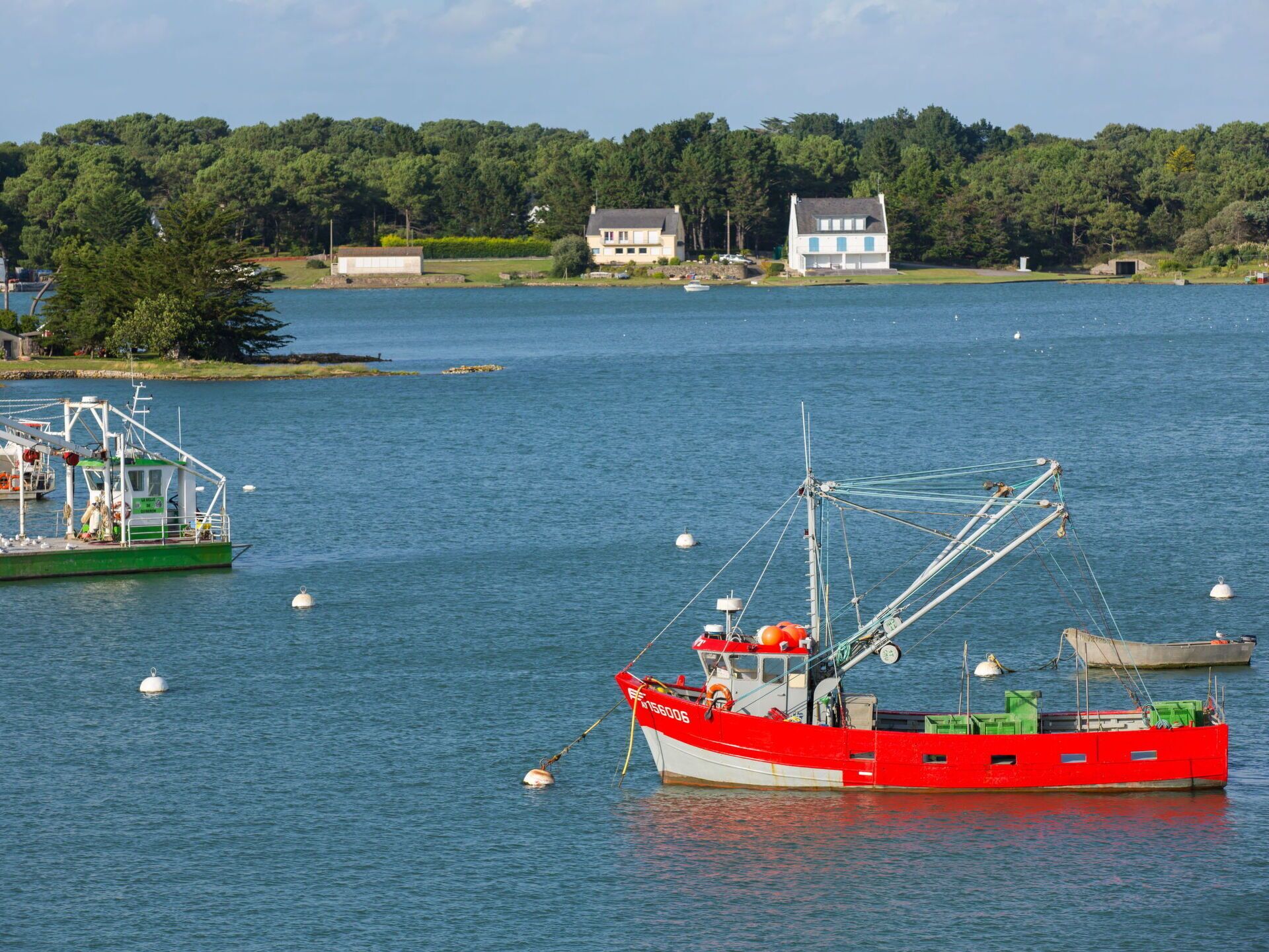 Um barco de pesca vermelho ancorado perto de um cais, com uma linha de costa arborizada ao fundo.