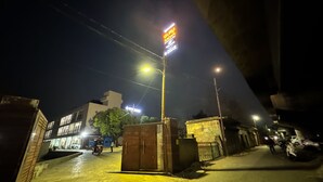 Escena nocturna de una calle con un cartel destacado que dice "Guru Sadan" y un edificio de varios pisos al fondo.