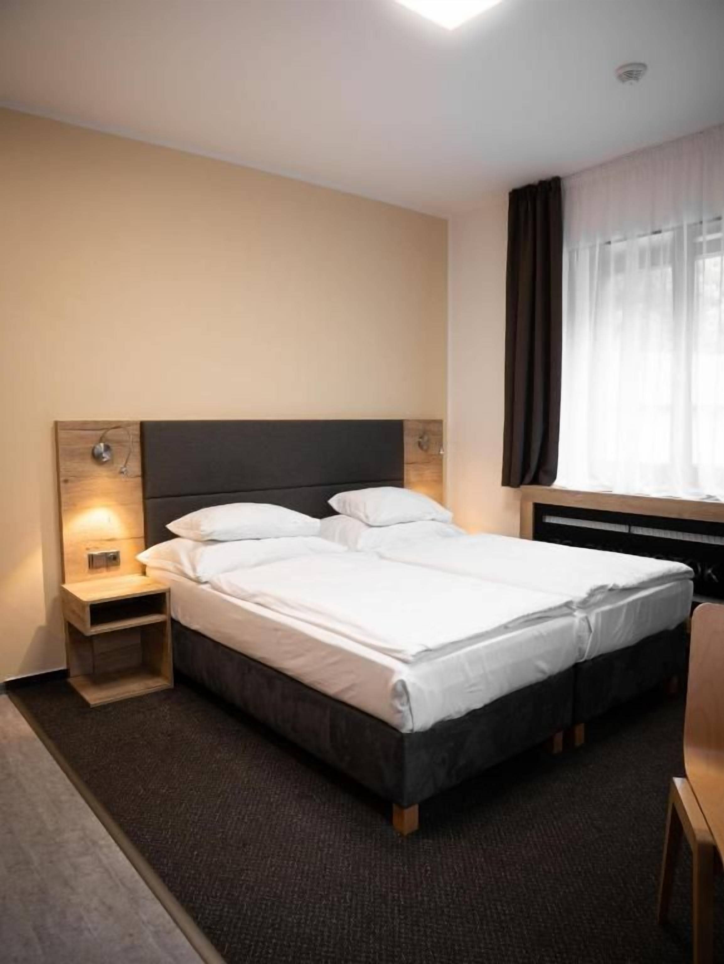Una habitación de hotel con cama, mesita de noche y ventana con cortinas.
